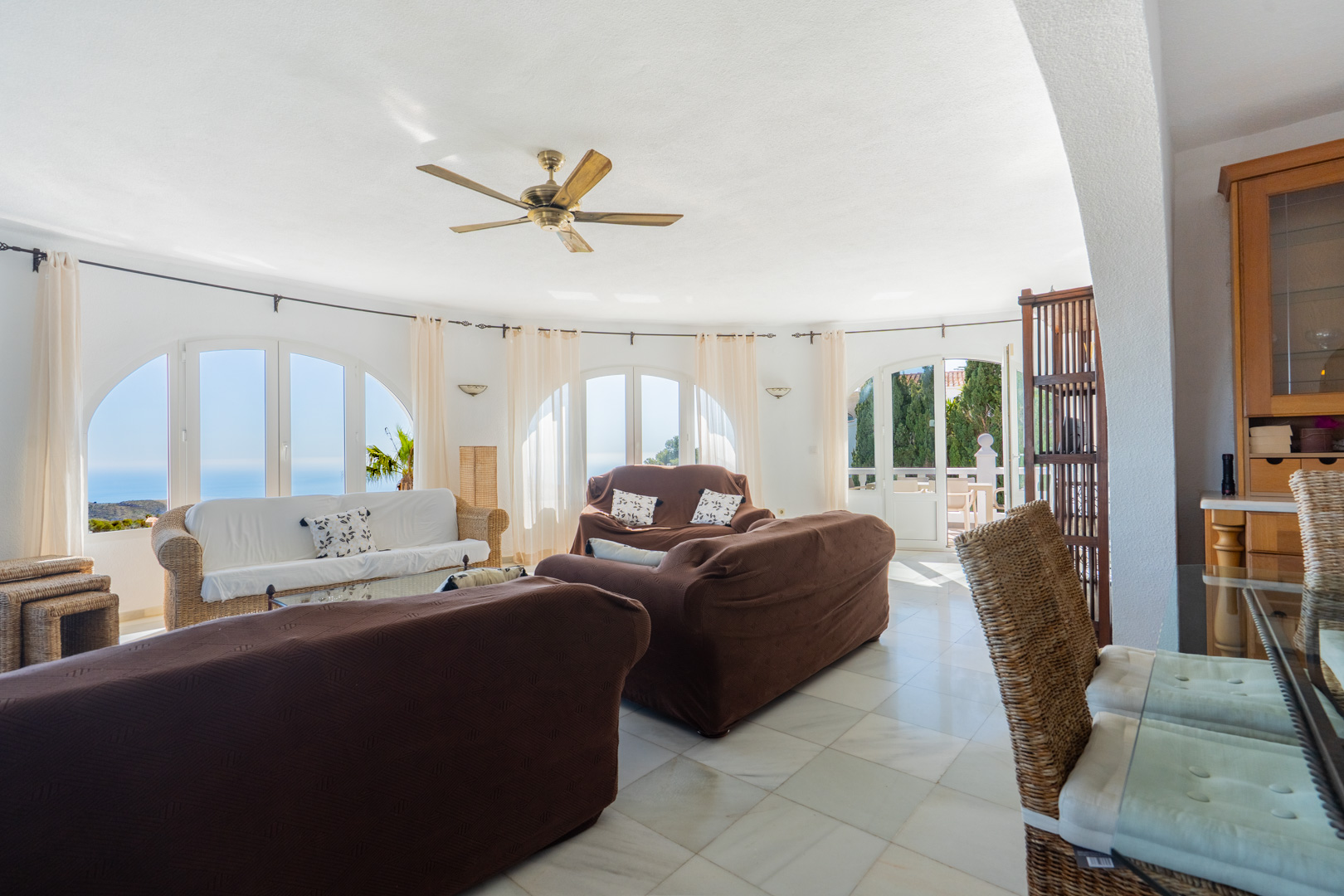 Salón circular con ventanales de arco y vistas al mar, suelos de mármol claro, ventilador de techo de madera y mobiliario con detalles en mimbre.
