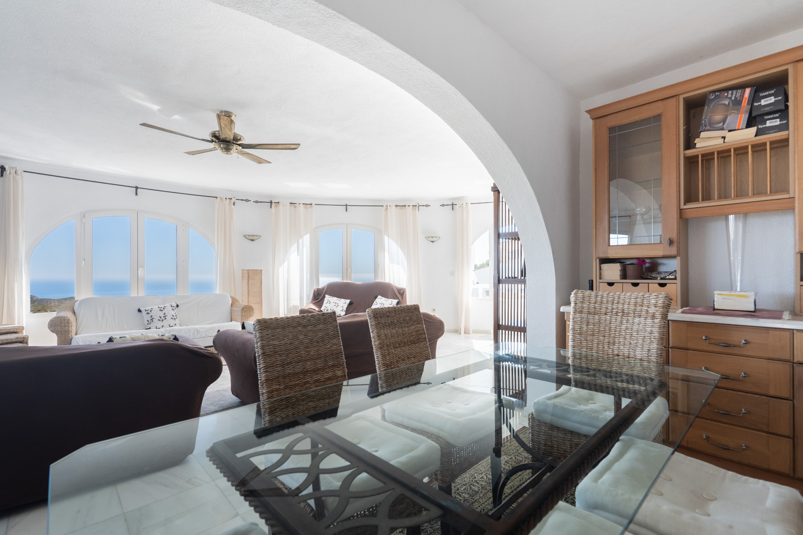 Salón-comedor de planta abierta con mesa de cristal, sillas de mimbre y muebles de madera integrados. Los ventanales arqueados ofrecen vistas al mar Mediterráneo.