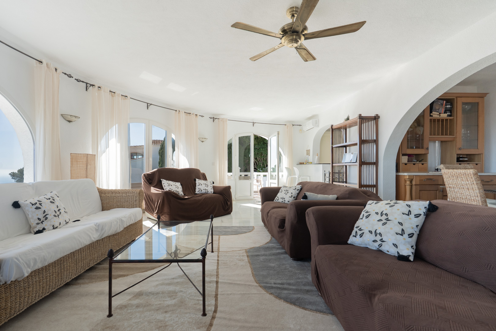 Salón luminoso con paredes curvas, ventanales de arco y acceso directo a la terraza. Incluye ventilador de techo de madera, suelos cerámicos y vista parcial a la cocina.