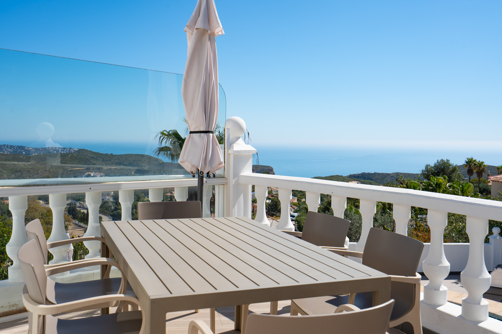 Terraza exterior con mesa de comedor y sillas, protegida por una balaustrada blanca y paneles de cristal, con vistas panorámicas al mar Mediterráneo y la costa.