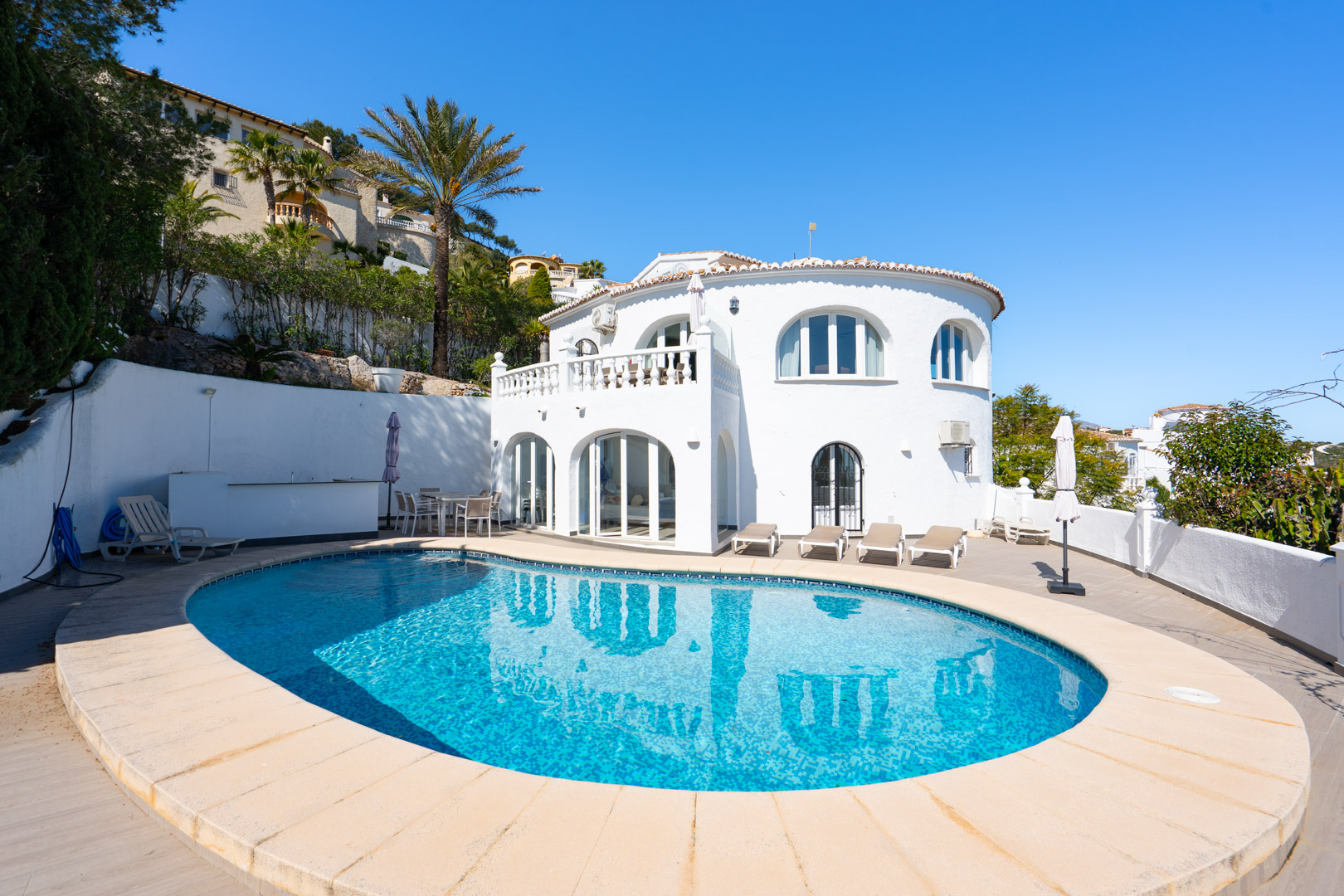 Vista exterior de la villa con piscina de forma ovalada, amplia terraza solárium y fachada blanca tradicional con arcos y naya acristalada.