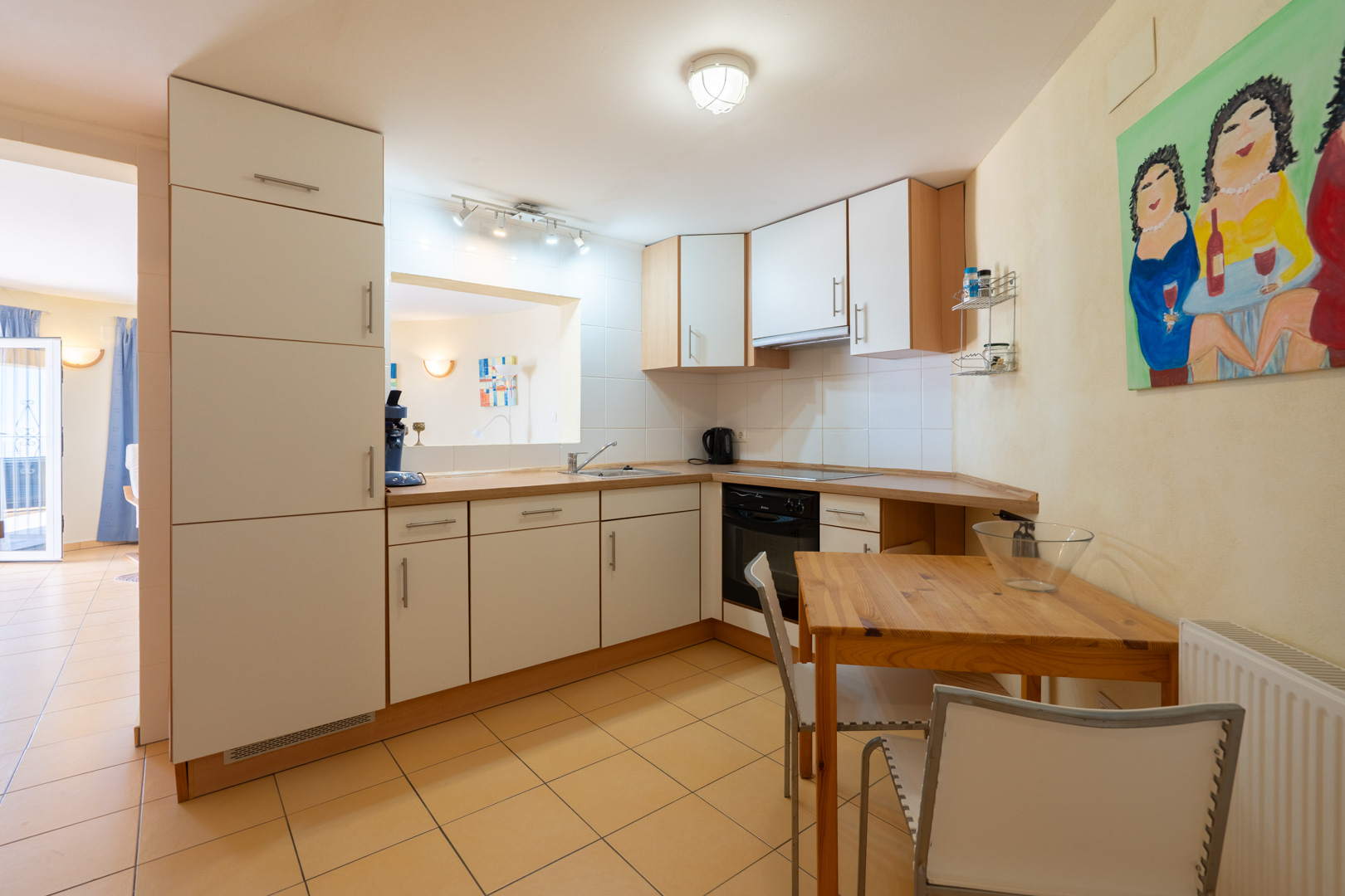 Cocina funcional con armarios blancos, encimera de madera y zona de comedor integrada. Incluye ventana pasaplatos y suelos cerámicos en tonos cálidos.