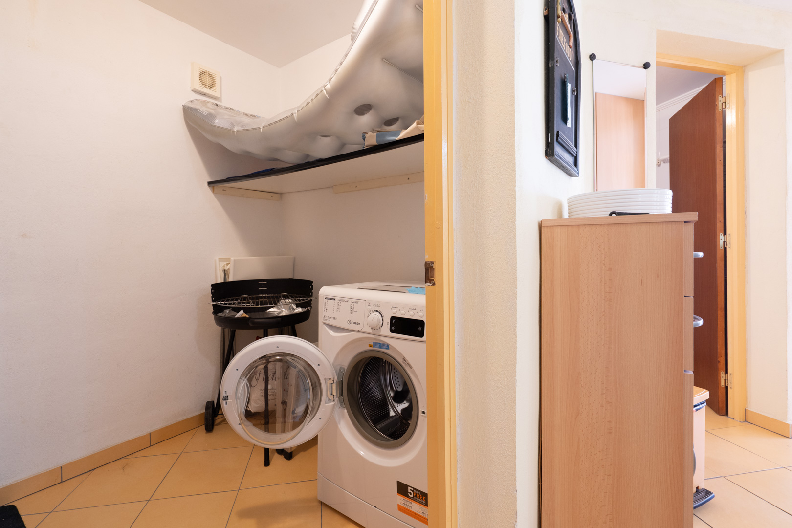 Cuarto de lavandería funcional con lavadora de carga frontal, estante de almacenamiento y suelos cerámicos, con acceso directo a otras estancias.