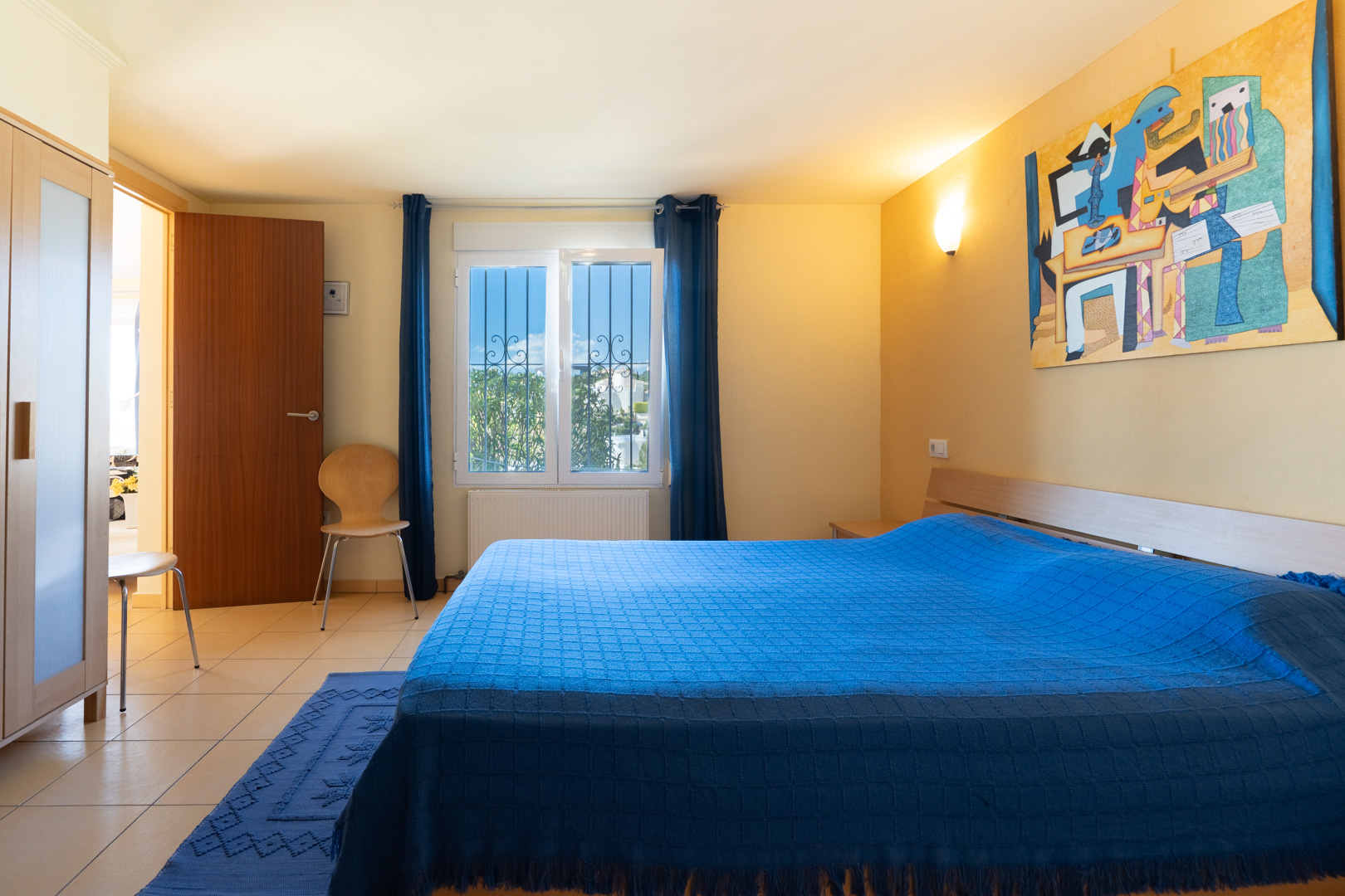 Dormitorio con cama doble, textiles azules, armario de madera y ventana con rejas. La estancia cuenta con suelos de gres y luz natural.