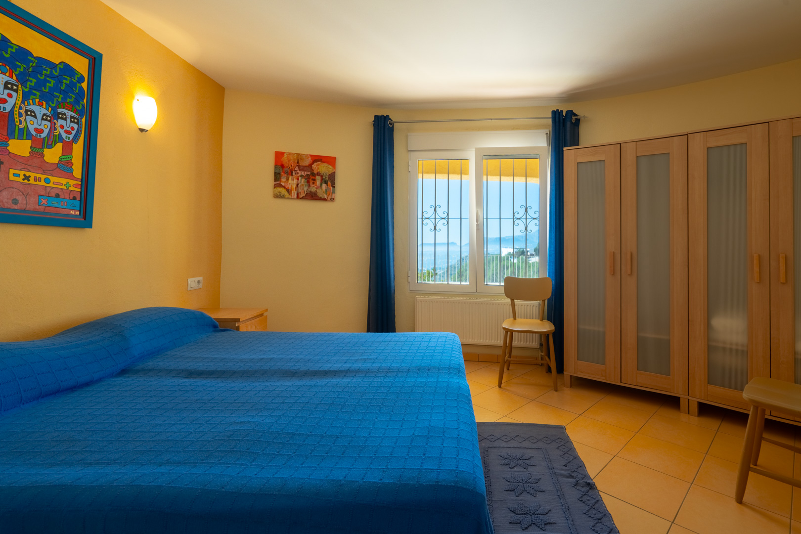 Dormitorio luminoso con cama doble, paredes ocres, armario de madera con paneles translúcidos y ventana con vistas panorámicas y radiador.