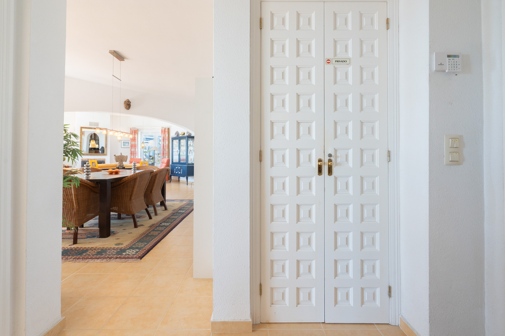 Entrada interior con puerta doble blanca panelada y vista al comedor principal con sillas de mimbre y suelos cerámicos.