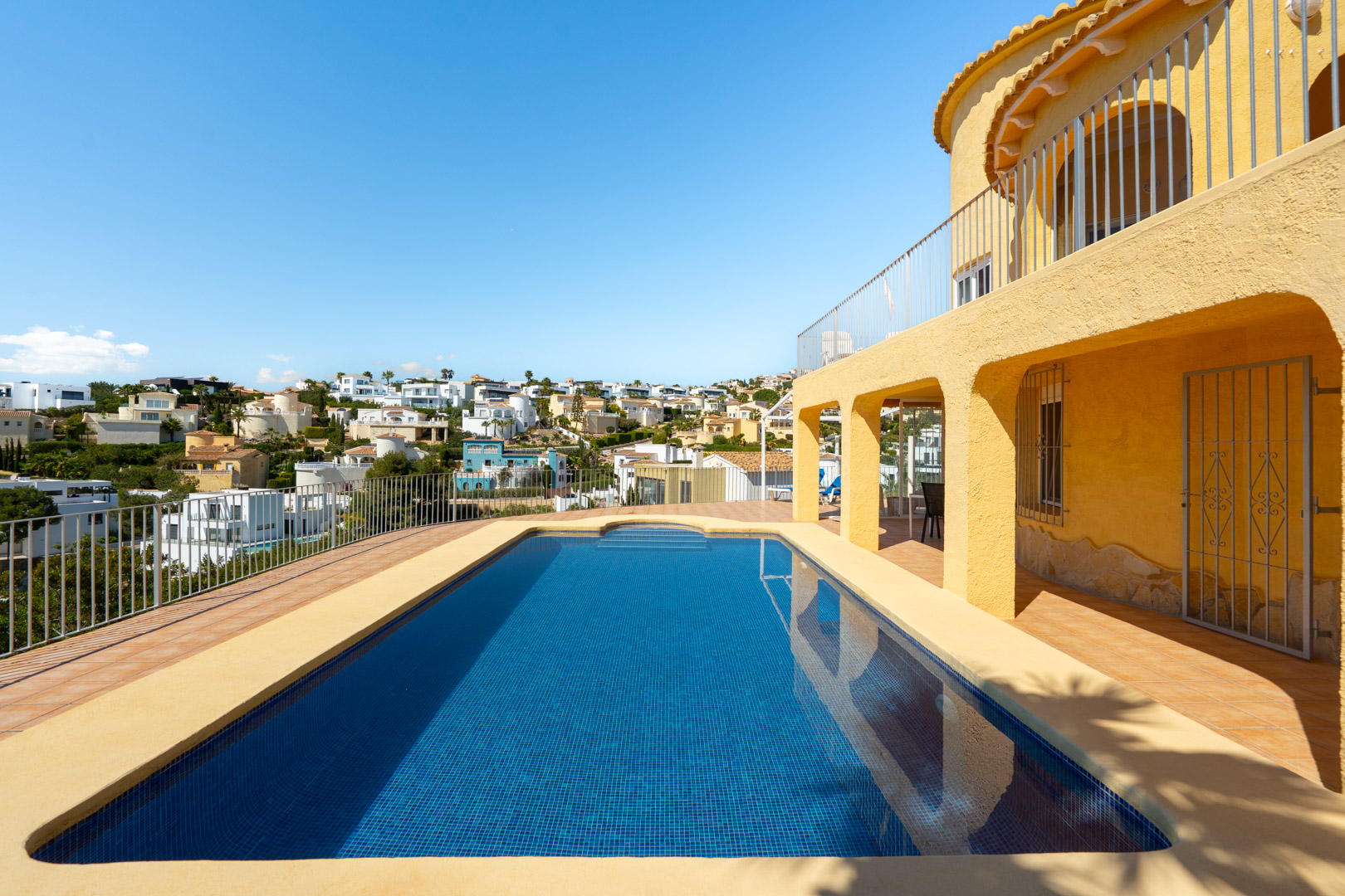 Piscina rectangular con gresite azul rodeada de una amplia terraza de baldosas cerámicas, junto a la fachada amarilla con arcos y vistas a la urbanización.