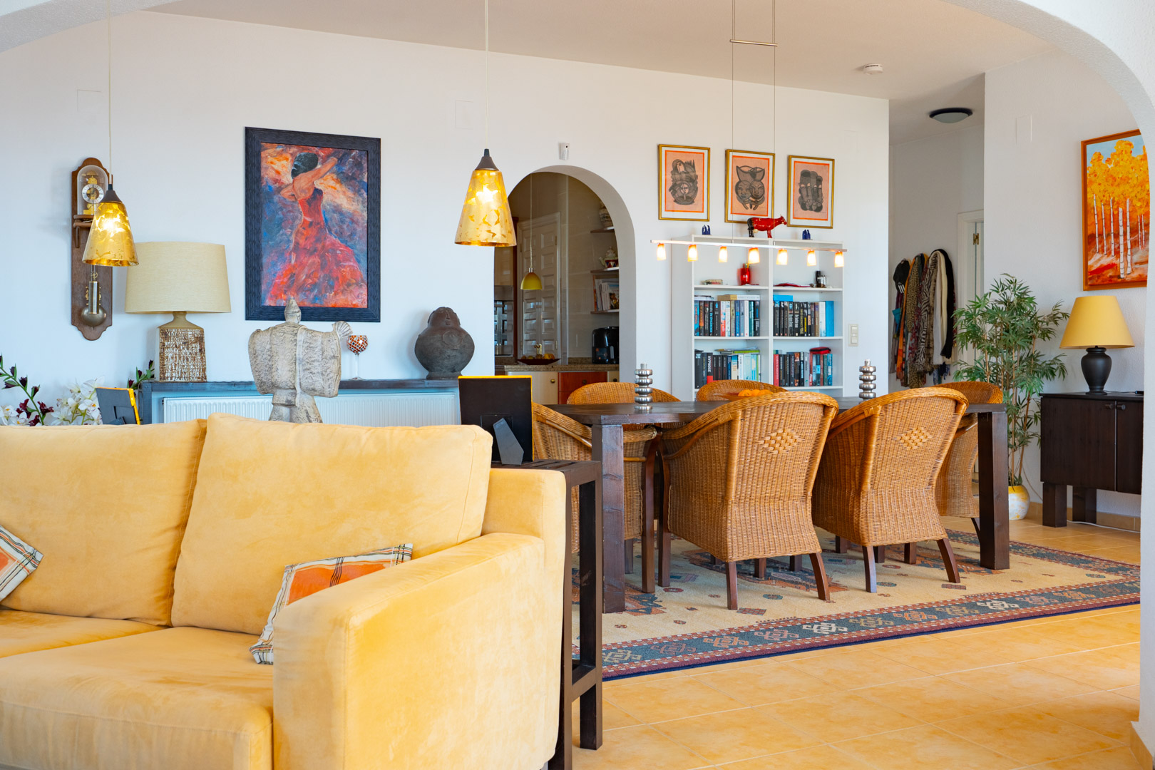 Salón-comedor de concepto abierto con sillas de mimbre, mesa de madera oscura y suelos cerámicos. La estancia destaca por su iluminación cálida y el acceso arqueado a la cocina.
