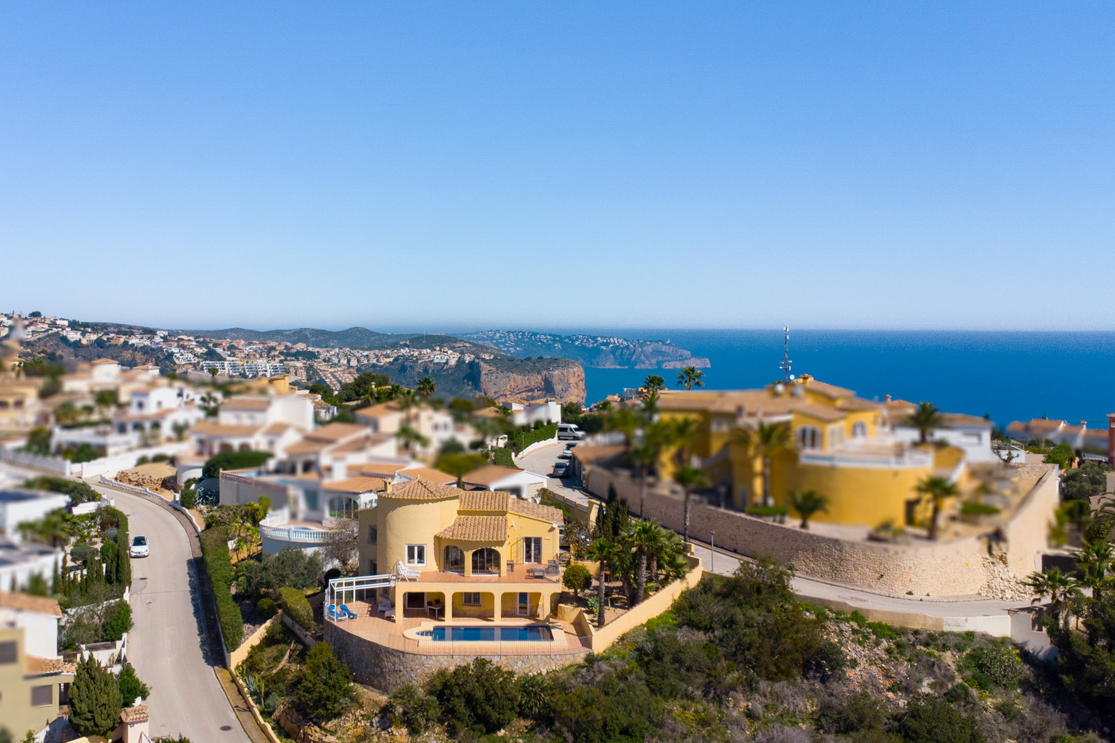 Vista aérea de la villa con fachada amarilla, destacando su piscina privada y terrazas con vistas panorámicas al mar Mediterráneo y al Cap d’Or en Moraira.