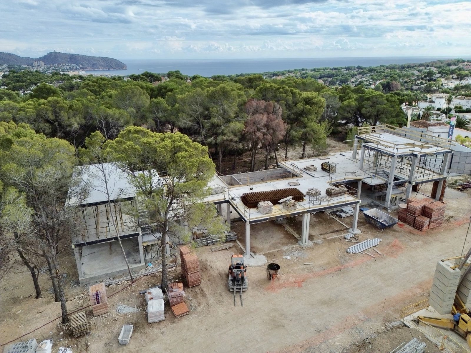 Vista aérea de la villa en fase de construcción, mostrando su estructura de hormigón y materiales de obra. Al fondo, se aprecian el mar Mediterráneo y el paisaje arbolado. Vista aérea de la villa en fase de construcción, mostrando su estructura de hormigón y materiales de obra. Al fondo, se aprecian el mar Mediterráneo y el paisaje arbolado.
