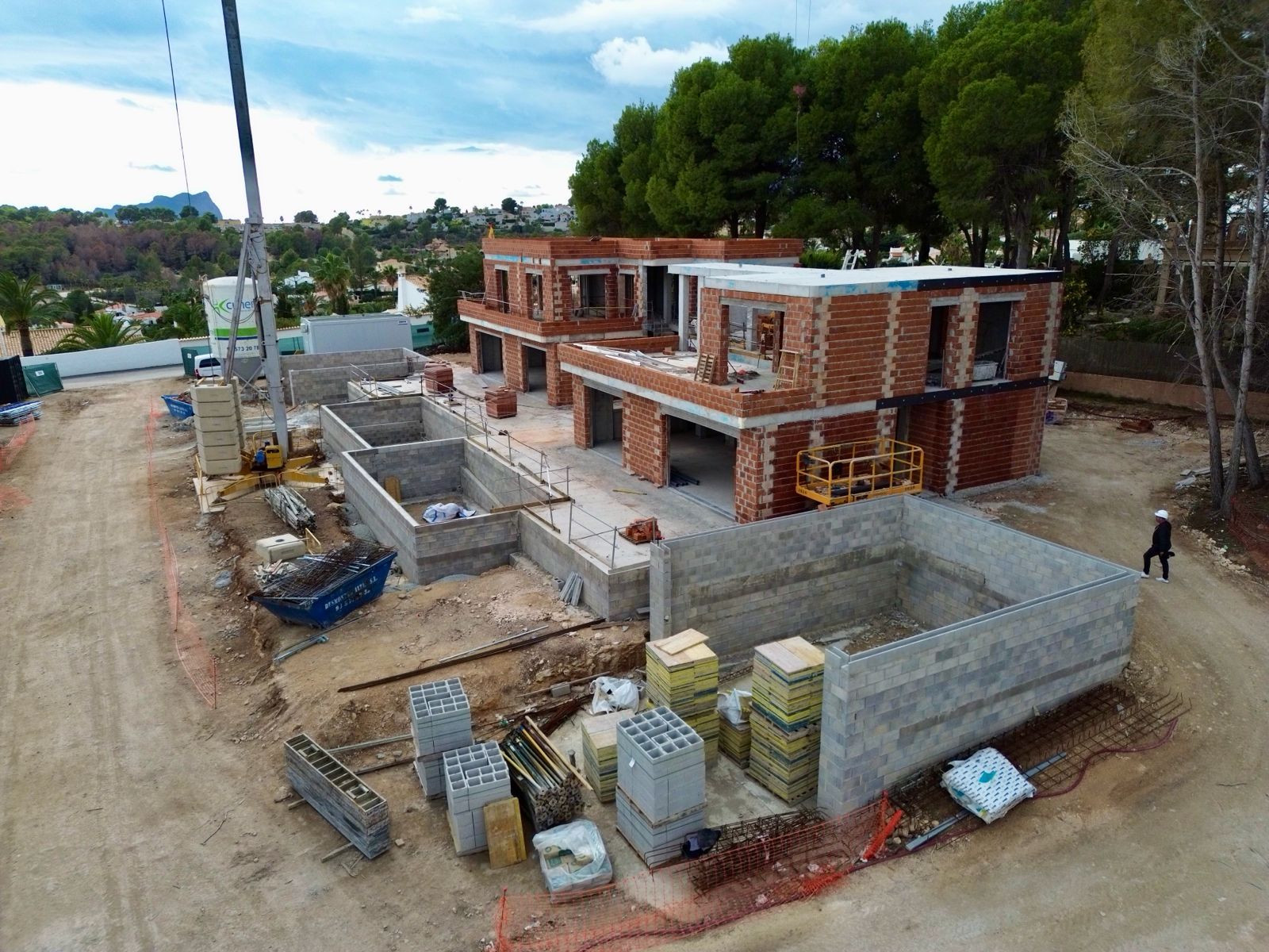 Vista aérea del sitio de construcción de la villa, mostrando múltiples estructuras de ladrillo en progreso, cimientos de hormigón excavados y materiales de construcción. Una grúa es visible, con colinas verdes distantes. Vista aérea del sitio de construcción de la villa, mostrando múltiples estructuras de ladrillo en progreso, cimientos de hormigón excavados y materiales de construcción. Una grúa es visible, con colinas verdes distantes.