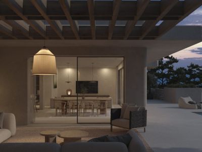 Acogedora zona de estar exterior bajo pérgola de madera, con sofás modernos y lámpara colgante, conectada a un comedor interior con vistas a la montaña al atardecer.