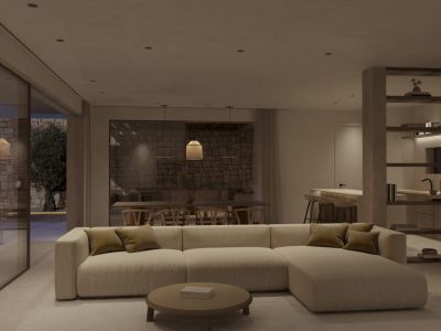 Salón-comedor de diseño abierto con sofá modular, mesa de centro redonda y acceso a terraza con muros de piedra y olivo. La cocina integrada se vislumbra al fondo.