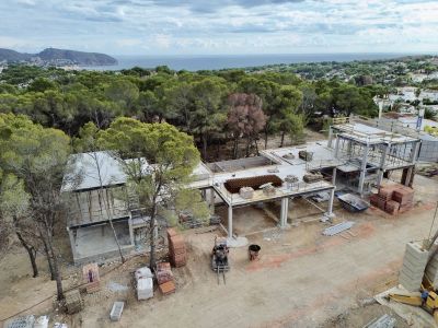 Vista aérea de la villa en fase de construcción, mostrando su estructura de hormigón y materiales de obra. Al fondo, se aprecian el mar Mediterráneo y el paisaje arbolado.
