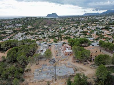 Vista aérea de un sitio de construcción de villas con cimientos de hormigón y paredes parcialmente levantadas, en una zona residencial con vistas lejanas al mar y la montaña.