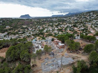 Vista aérea de villas en construcción en una zona residencial de Moraira, con grúas y cimientos visibles, enmarcada por el mar y el Peñón de Ifach a lo lejos.