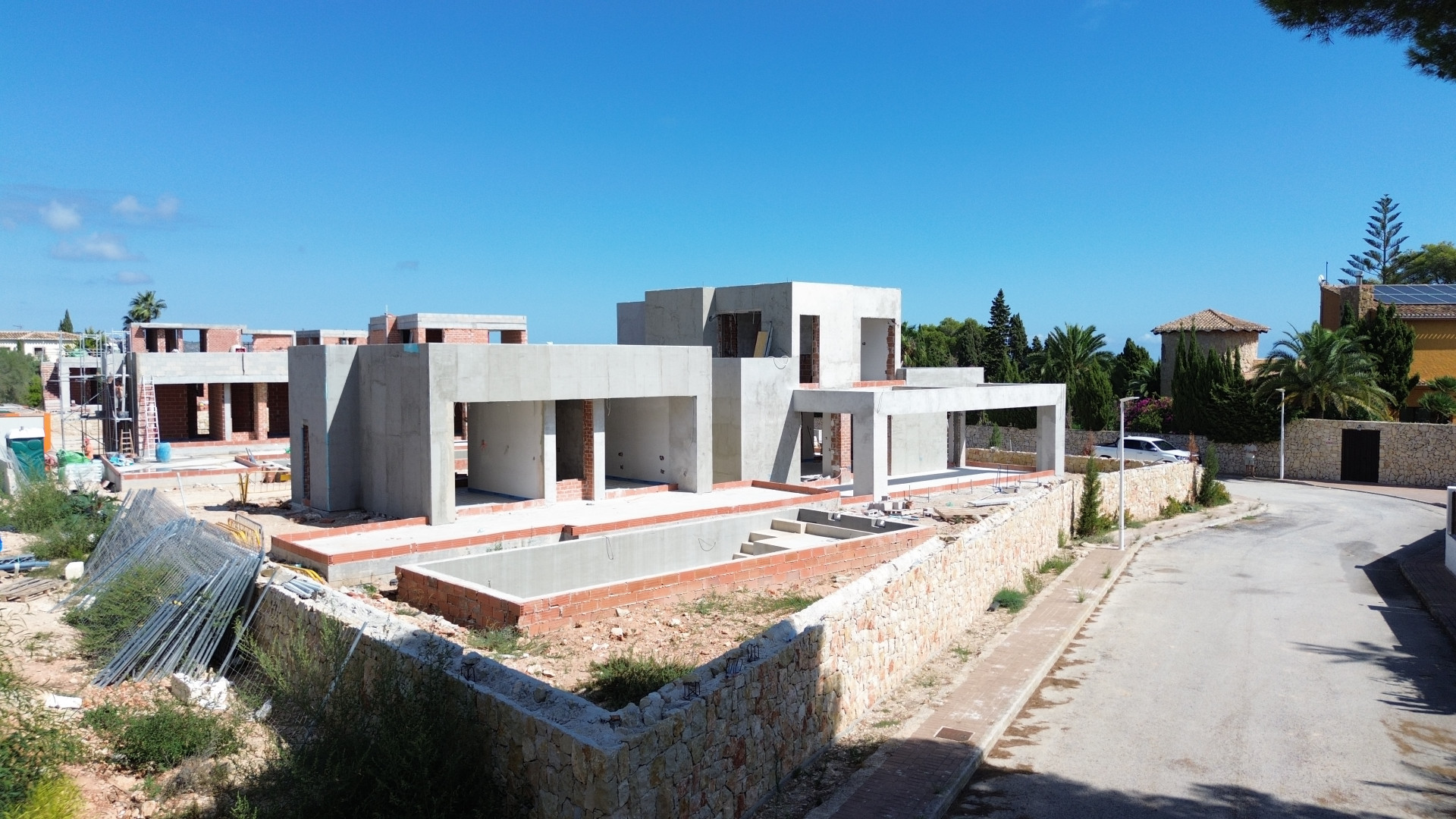 Villa moderna en construcción, mostrando su estructura de hormigón y la excavación para la futura piscina privada. Muros de piedra definen el límite de la propiedad junto a la tranquila calle residencial. Villa moderna en construcción, mostrando su estructura de hormigón y la excavación para la futura piscina privada. Muros de piedra definen el límite de la propiedad junto a la tranquila calle residencial.
