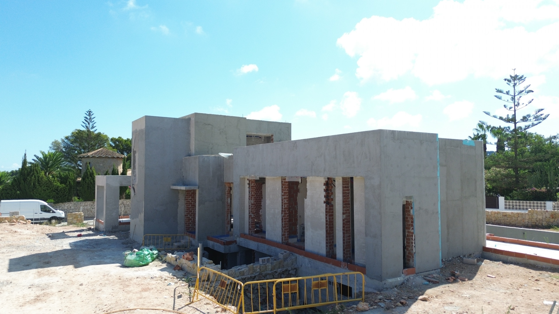 Vista exterior de la villa en construcción, mostrando la fachada enlucida en gris, pilares de ladrillo expuesto y el terreno con barreras de obra. Vegetación y un edificio tradicional se aprecian al fondo. Vista exterior de la villa en construcción, mostrando la fachada enlucida en gris, pilares de ladrillo expuesto y el terreno con barreras de obra. Vegetación y un edificio tradicional se aprecian al fondo.