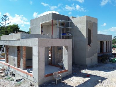 Estructura de hormigón de una villa moderna en construcción, con grandes aberturas para ventanales y andamios visibles. El terreno de obra muestra materiales de construcción bajo un cielo azul.