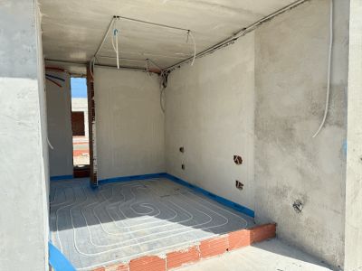 Habitación en fase de construcción con instalación de suelo radiante visible y paredes de hormigón. Se aprecian conductos eléctricos y una entrada a otra zona sin terminar.