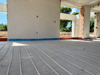 Instalación de tuberías de suelo radiante sobre base de hormigón, con paredes parcialmente enlucidas y pilares de ladrillo visto, en fase de construcción. Luz natural desde el exterior.