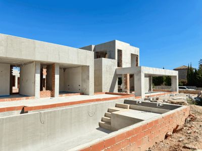 Villa en construcción, mostrando su estructura de hormigón y la piscina privada en obra con escaleras bajo un cielo azul claro.