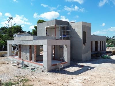Villa en fase de construcción con estructura de hormigón y ladrillo visto, andamios en el nivel superior y terreno de obra circundante bajo un cielo azul claro.