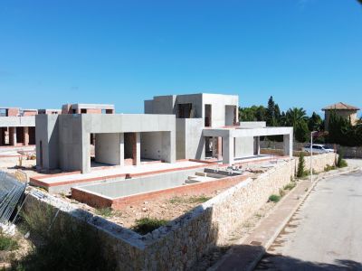 Villa moderna en construcción, mostrando su estructura de hormigón y la excavación para la futura piscina privada. Muros de piedra definen el límite de la propiedad junto a la tranquila calle residencial.
