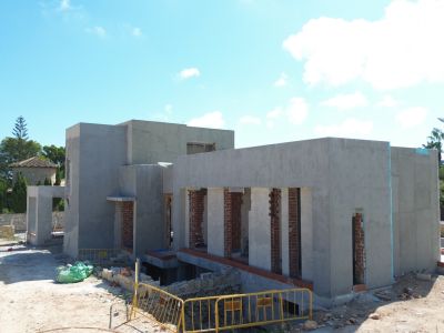 Vista exterior de la villa en construcción, mostrando la fachada enlucida en gris, pilares de ladrillo expuesto y el terreno con barreras de obra. Vegetación y un edificio tradicional se aprecian al fondo.