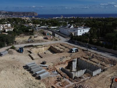 Vista aérea del solar en construcción con cimientos de hormigón y excavaciones. Al fondo, el paisaje de Jávea con el mar Mediterráneo y el Cabo de San Antonio.