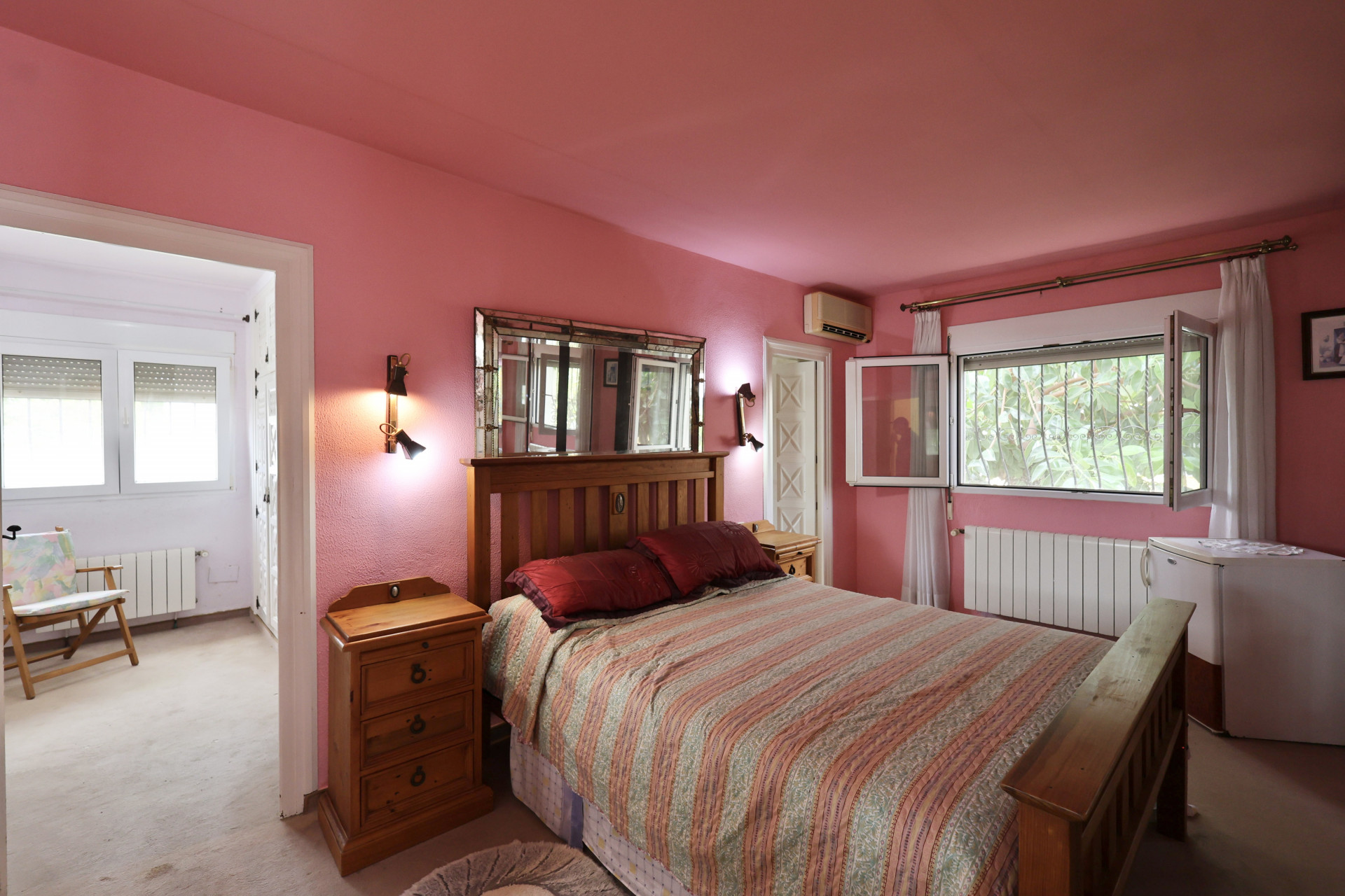 Dormitorio con paredes rosas, cama de madera y espejo con apliques. Requiere reforma.