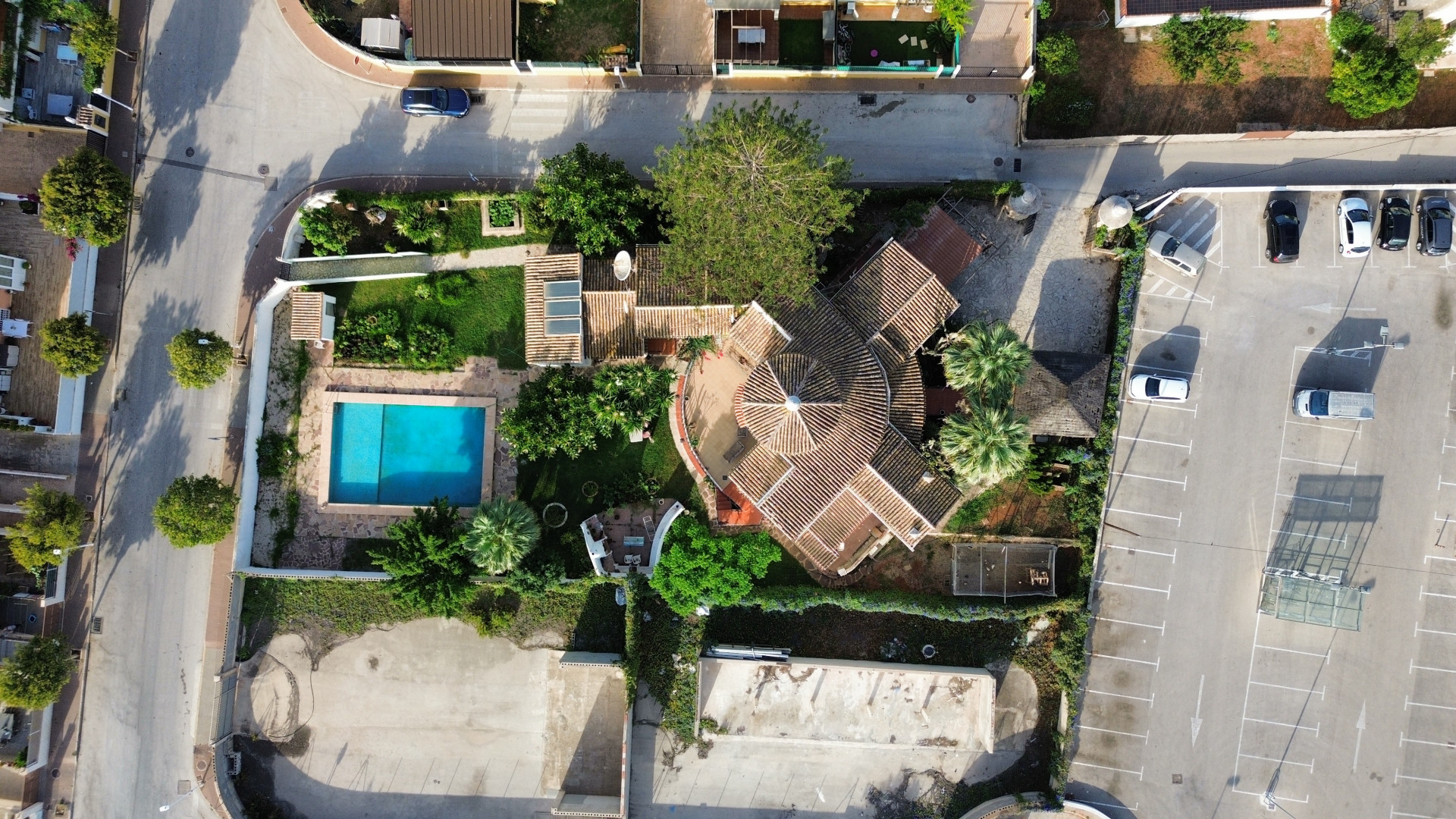 Vista aérea de la villa con piscina y tejado de tejas, rodeada de vegetación y aparcamiento adyacente.