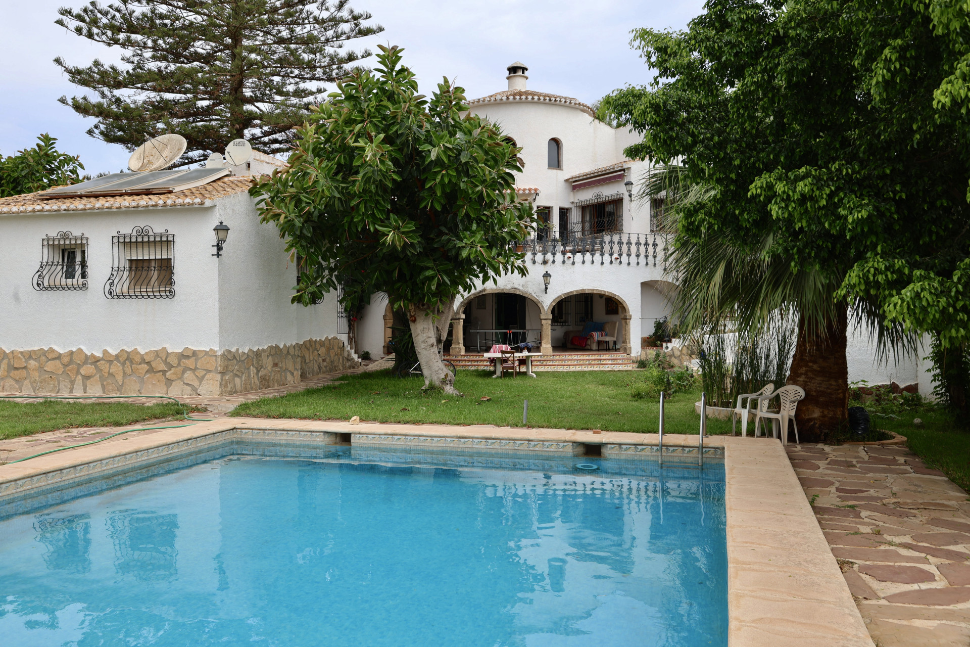 Vista de la piscina azul turquesa y la fachada blanca de la villa con detalles de piedra y jardín.