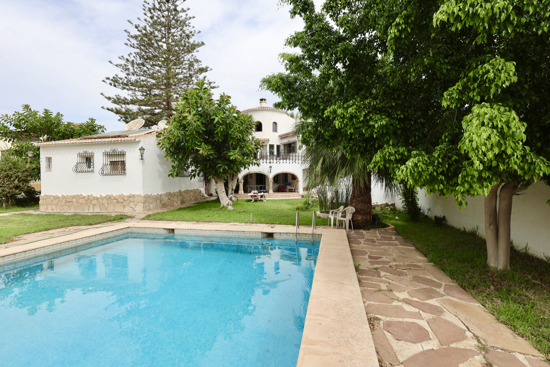 Vista de la piscina turquesa con borde de piedra, rodeada de césped y vegetación exuberante.