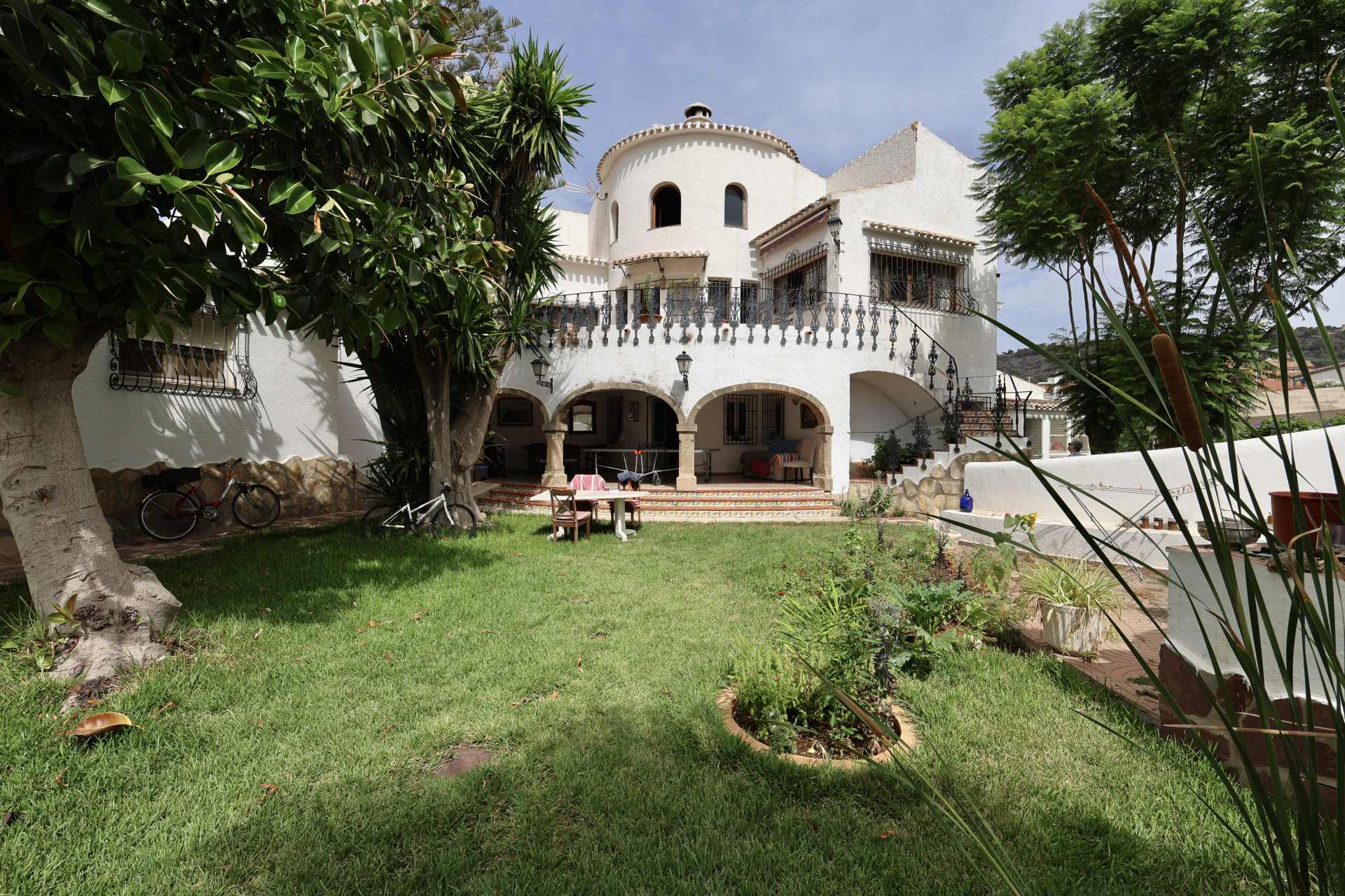 Vista del jardín con césped, la fachada blanca de la villa con arcos y una mesa de comedor.
