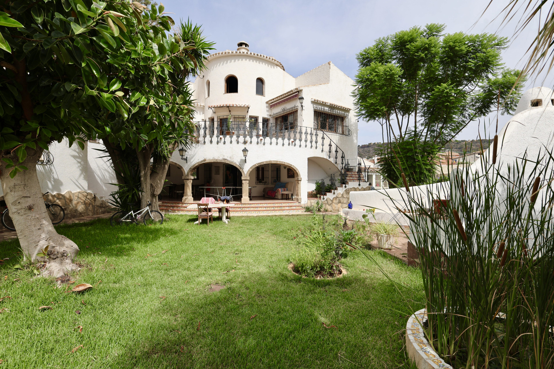 Vista del jardín delantero con césped, vegetación y fachada blanca de la villa de Jávea.