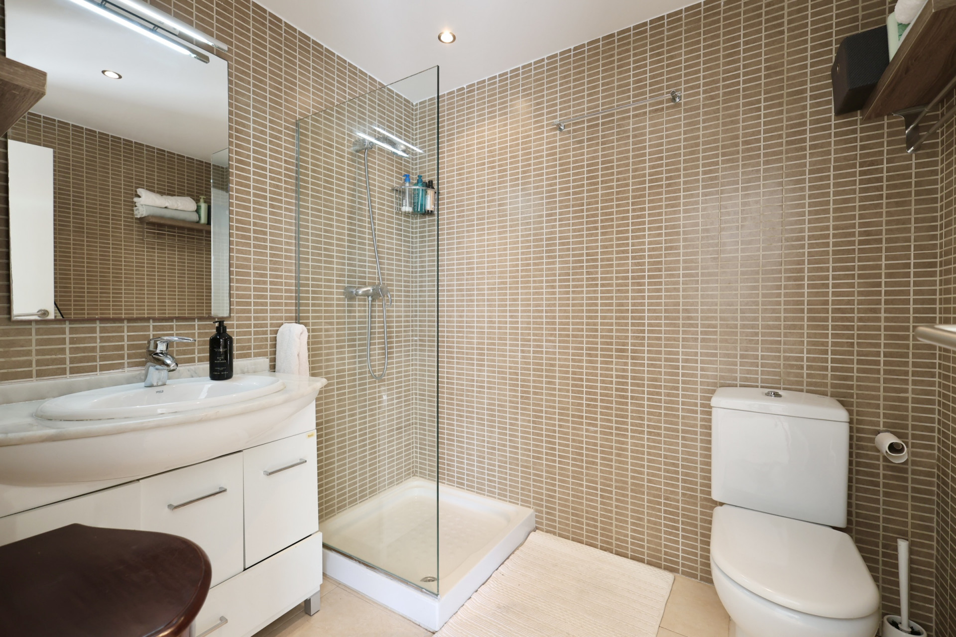 Cuarto de baño con azulejos color arena, plato de ducha con mampara de cristal y lavabo con encimera de mármol.