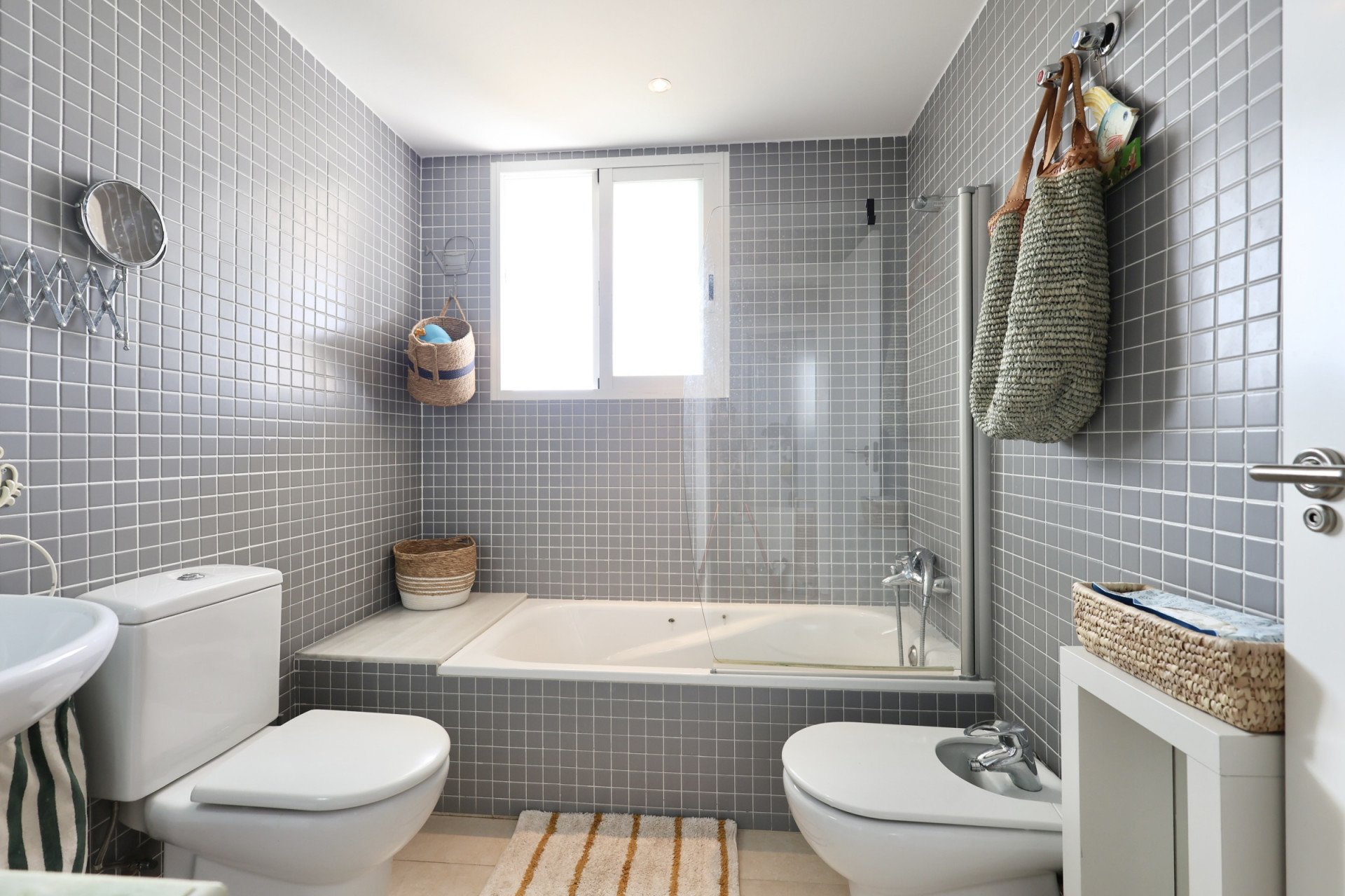 Cuarto de baño con azulejos grises, bañera con mampara y elementos decorativos de mimbre.