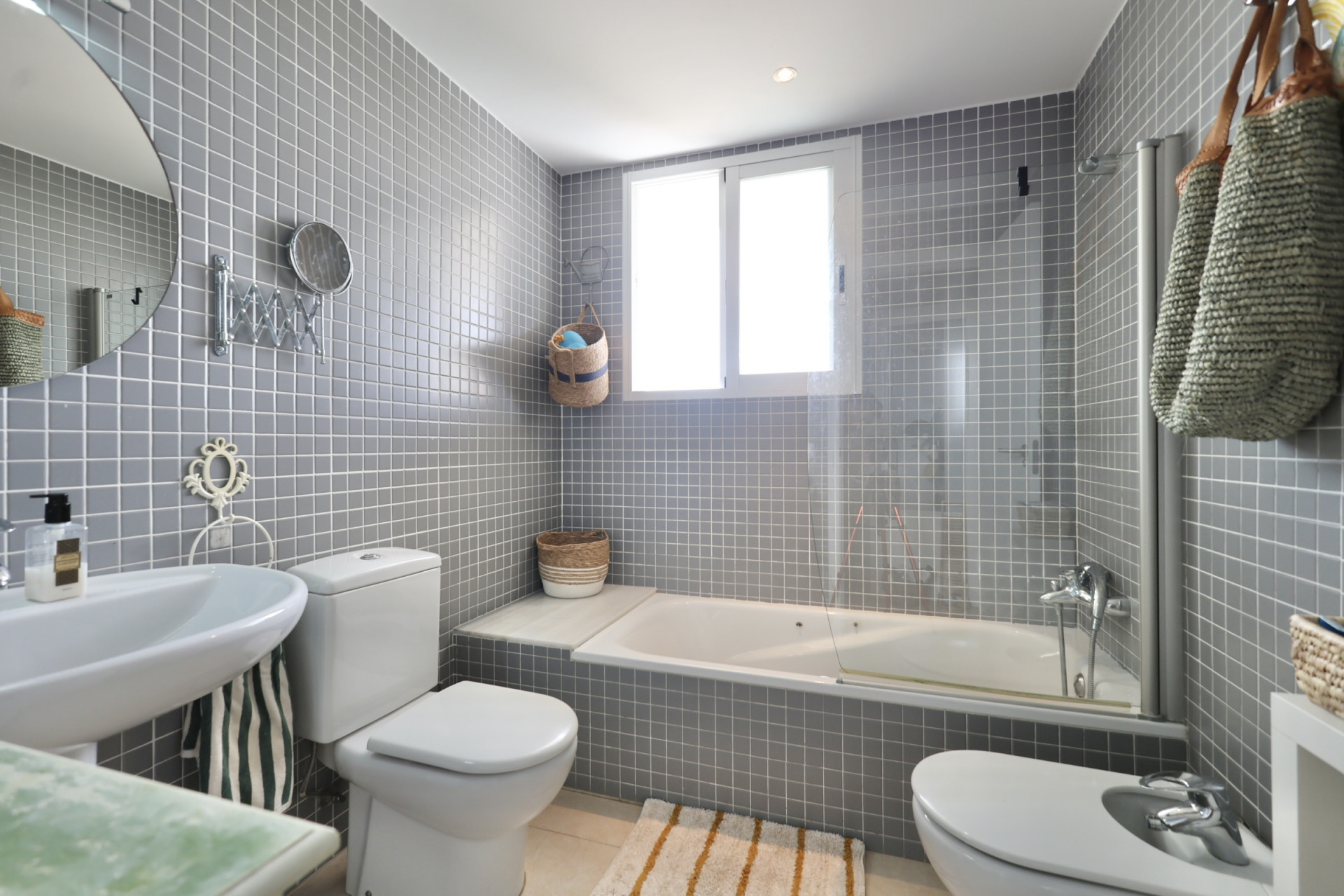 Cuarto de baño con azulejos grises, lavabo de pedestal, inodoro y bidé. Una bañera blanca se encuentra debajo de una ventana.