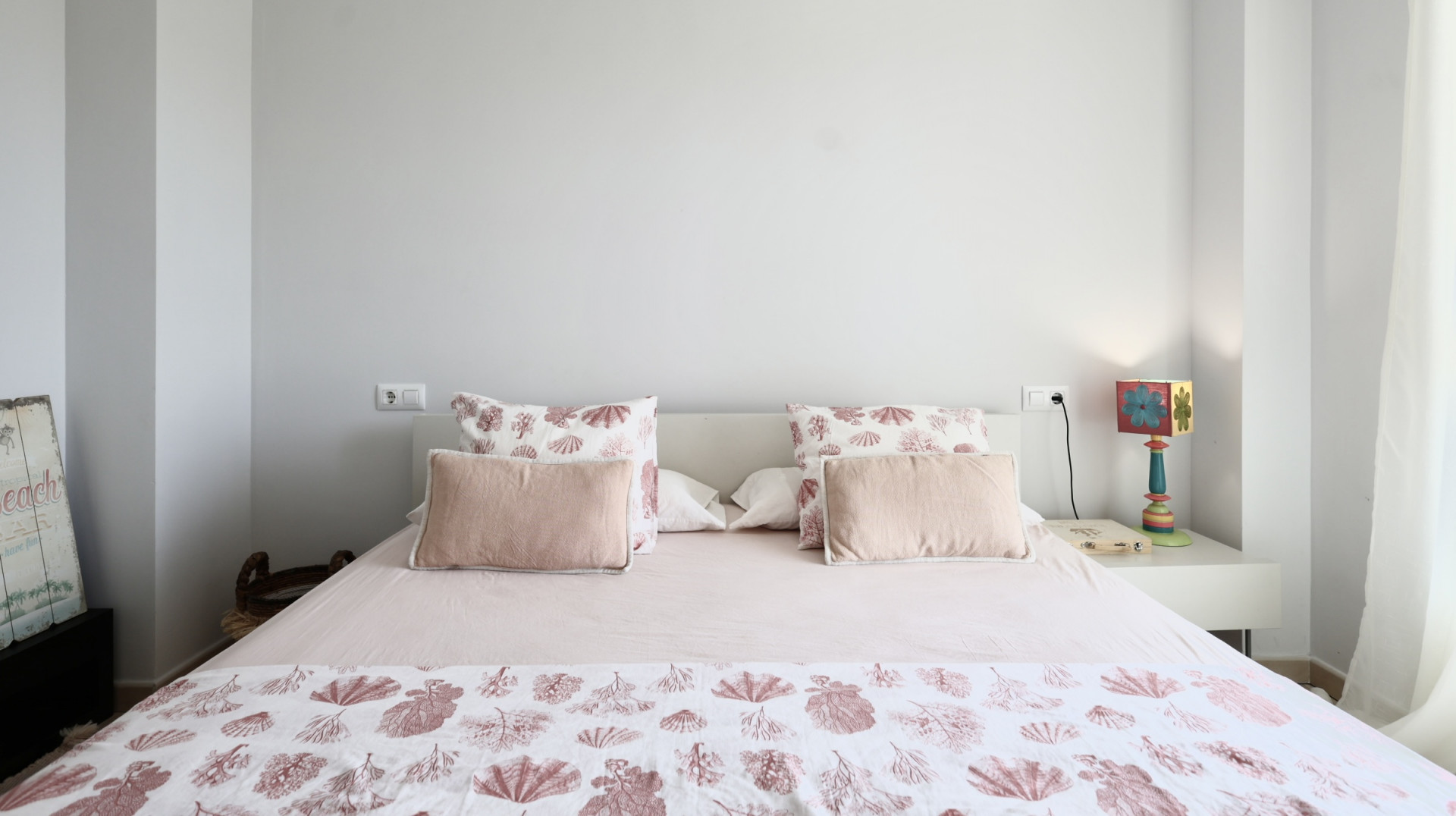 Dormitorio luminoso con cama doble, ropa de cama con motivos marinos y mesita de noche blanca con lámpara decorativa.