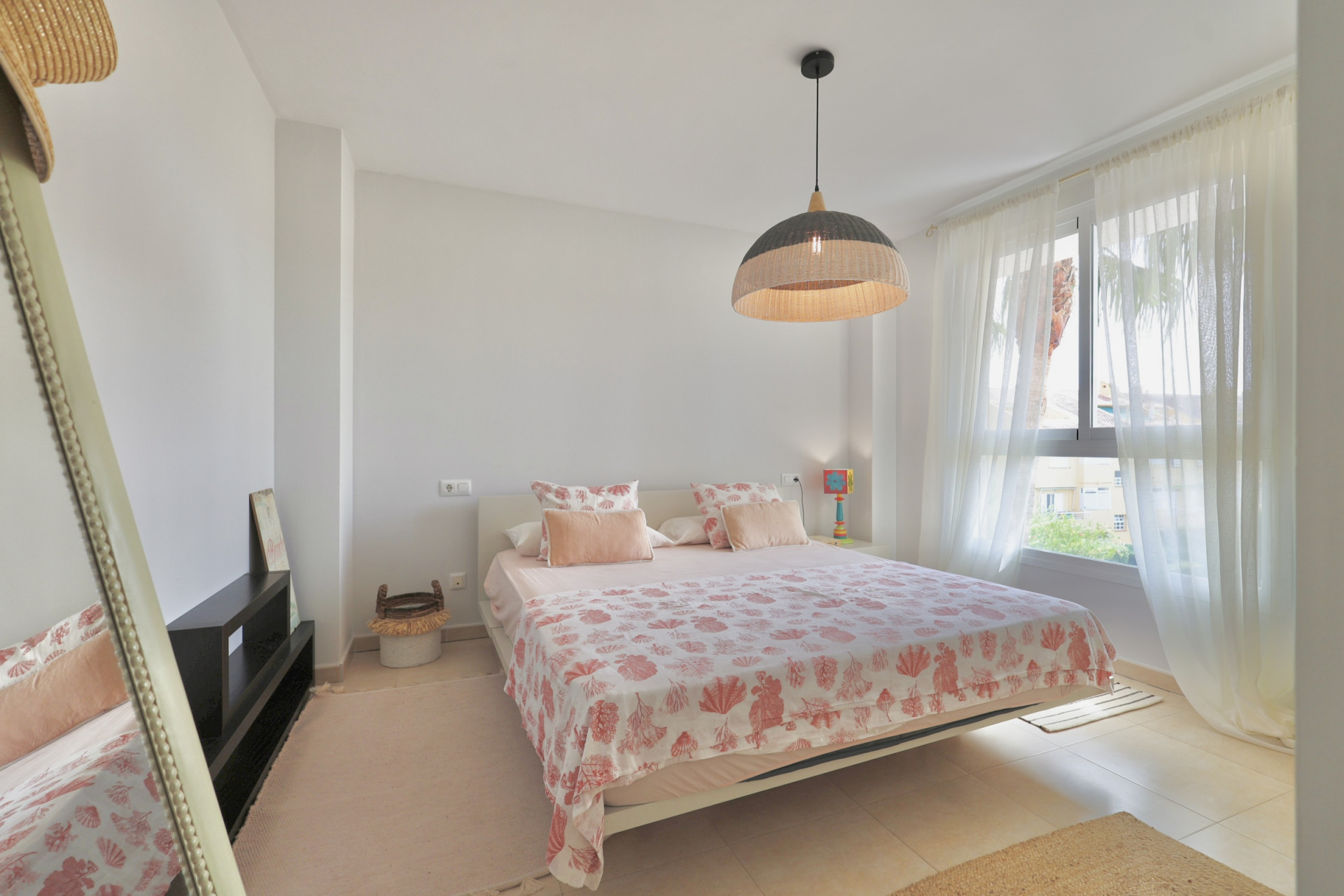 Dormitorio luminoso con cama doble, ropa de cama estampada y lámpara de mimbre colgante.