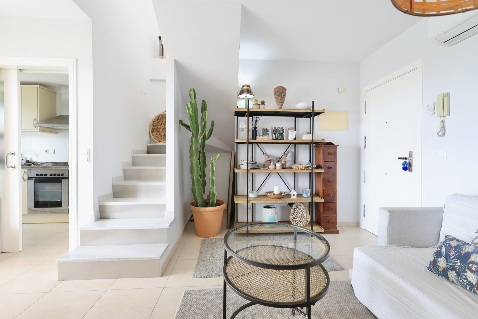 Interior luminoso con escalera blanca, estantería de madera y metal, y mesa de centro de ratán.
