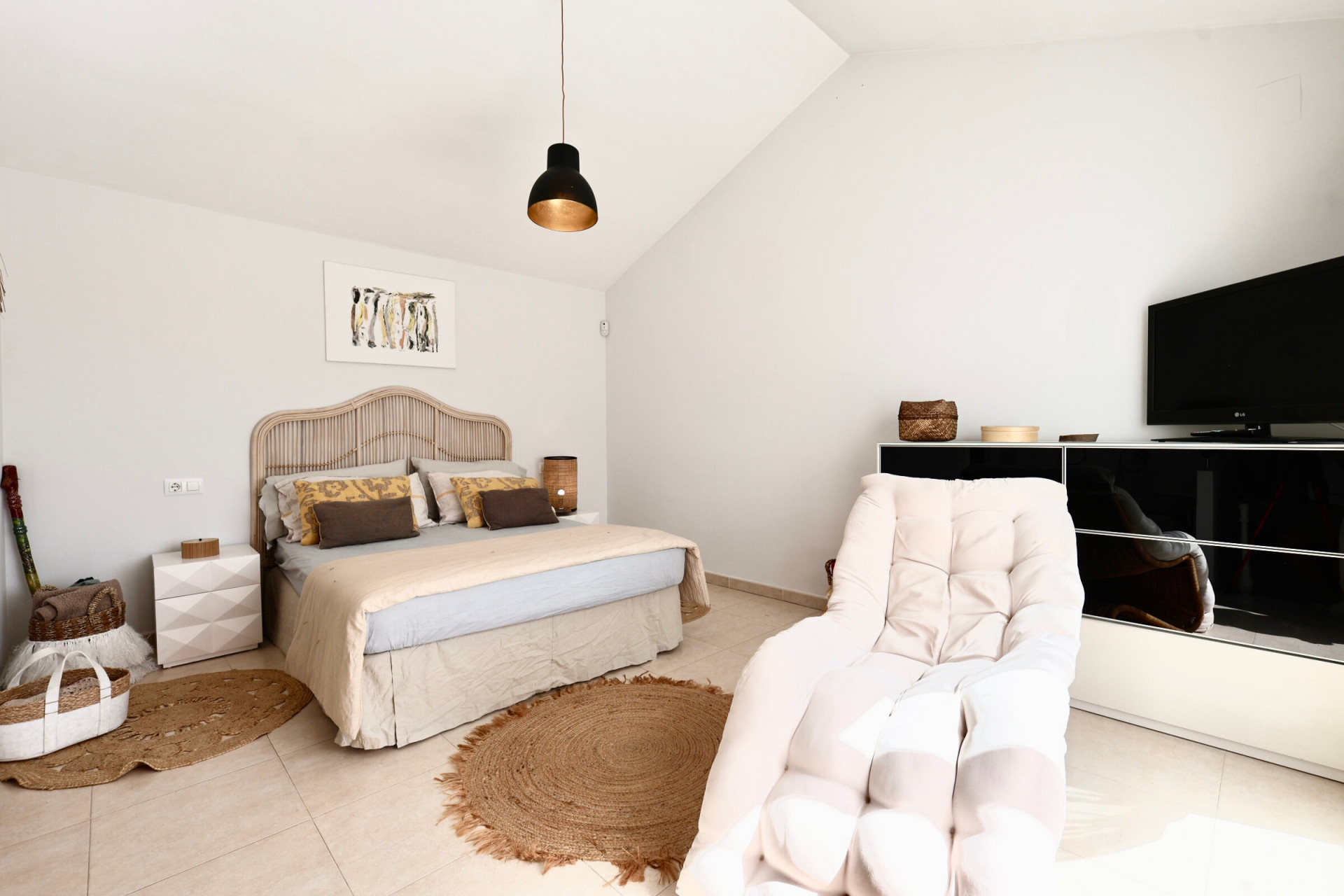 Luminoso dormitorio con cama de ratán, alfombra de yute y sillón junto a mueble de TV negro.