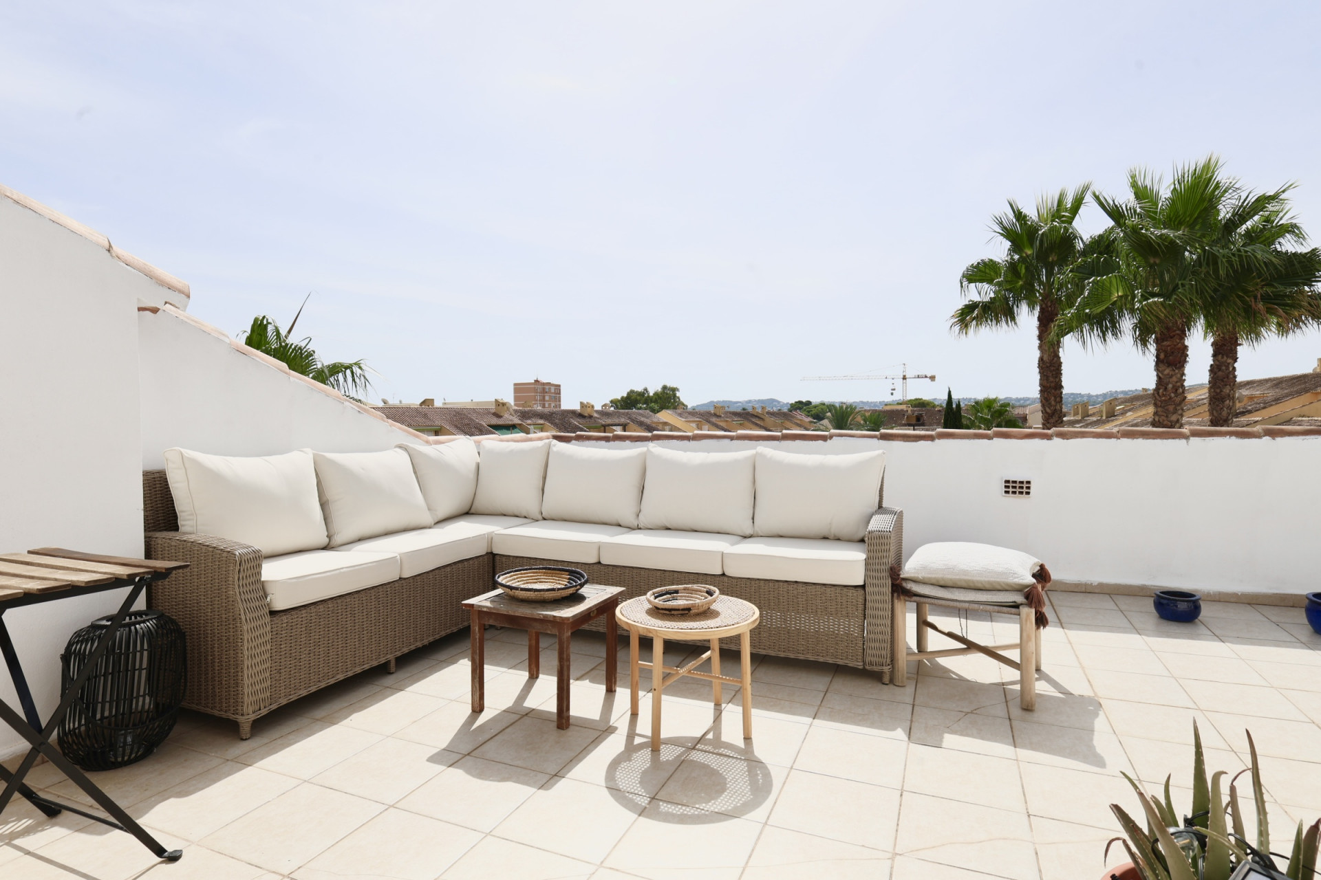 Terraza con sofá esquinero de mimbre y cojines blancos, con vistas a las palmeras y tejados.