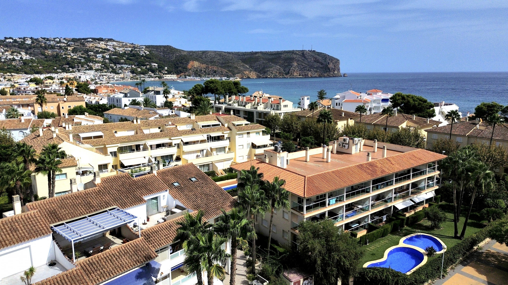 Vista aérea de la urbanización con piscina y tejados de tejas rojas, con el mar y la costa al fondo.