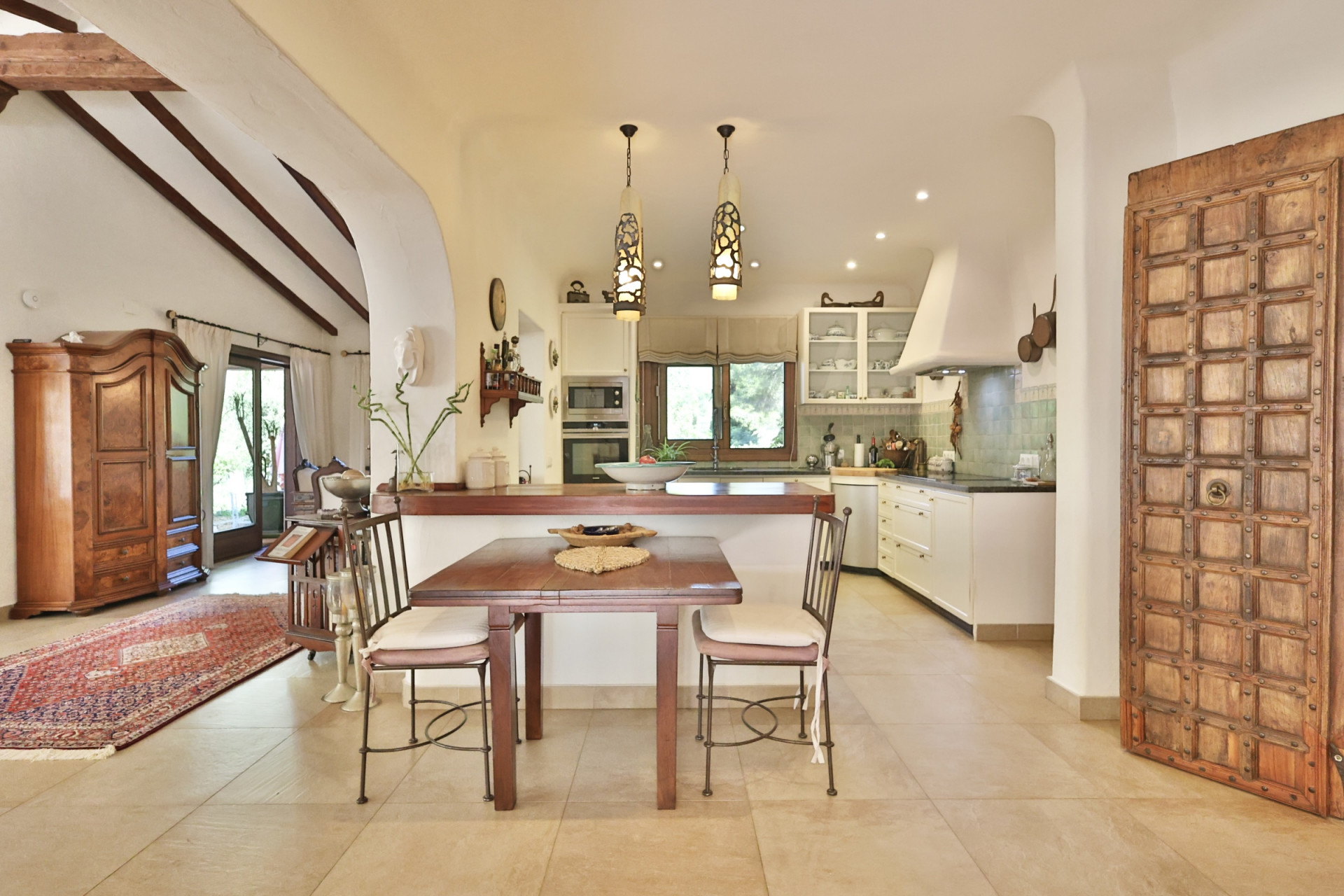 Cocina abierta con encimeras oscuras, azulejos verdes y electrodomésticos integrados. Mesa de comedor de madera junto a una puerta decorativa y un salón con vigas vistas. Cocina abierta con encimeras oscuras, azulejos verdes y electrodomésticos integrados. Mesa de comedor de madera junto a una puerta decorativa y un salón con vigas vistas.
