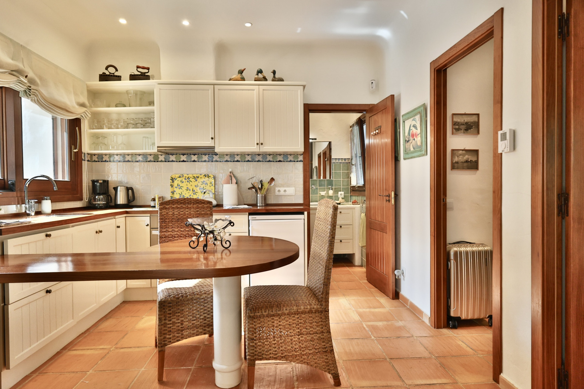 Cocina funcional con encimeras de madera, armarios blancos y suelo de terracota. Incluye una mesa redonda con sillas de mimbre y acceso a un baño con azulejos verdes. Cocina funcional con encimeras de madera, armarios blancos y suelo de terracota. Incluye una mesa redonda con sillas de mimbre y acceso a un baño con azulejos verdes.