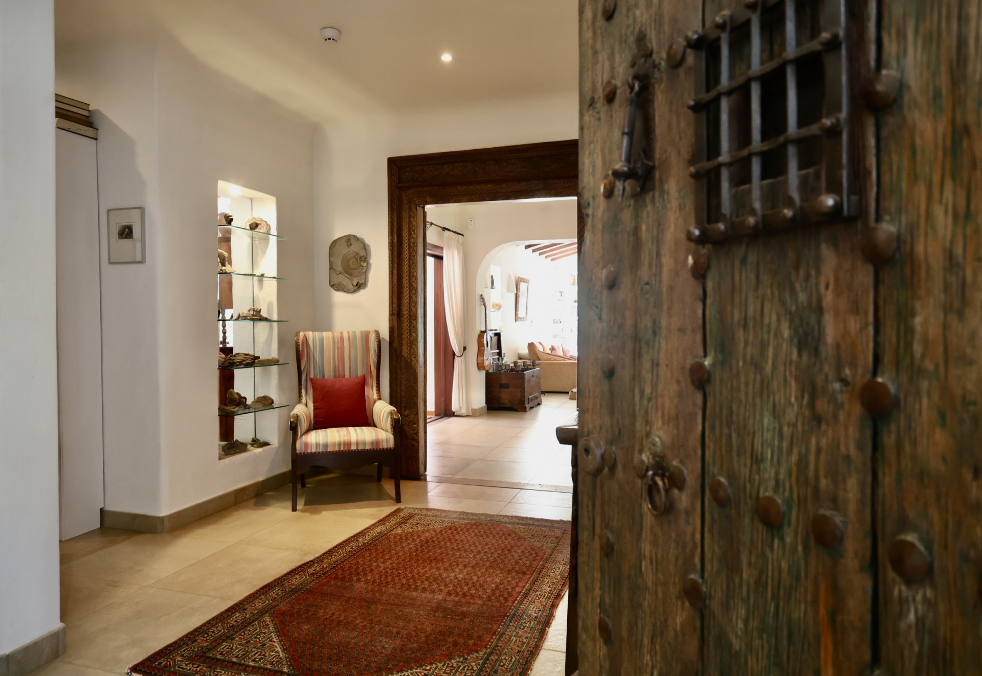 La rústica puerta de entrada de madera con herrajes metálicos y reja se abre a un luminoso recibidor. Destacan un sillón a rayas, una hornacina con estantes de cristal y una alfombra roja sobre suelo de baldosas. La rústica puerta de entrada de madera con herrajes metálicos y reja se abre a un luminoso recibidor. Destacan un sillón a rayas, una hornacina con estantes de cristal y una alfombra roja sobre suelo de baldosas.