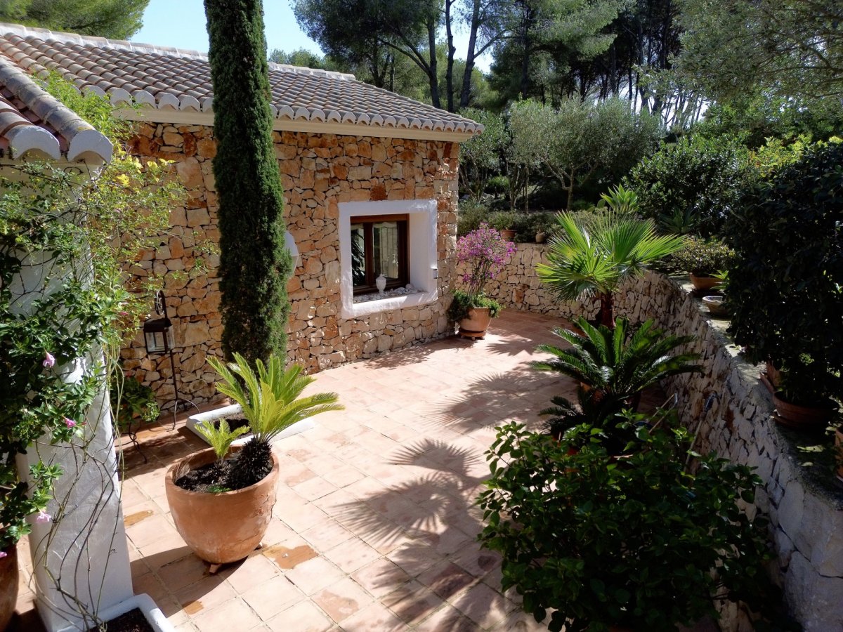Patio exterior con baldosas de terracota junto a una edificación de fachada de piedra. Incluye un ciprés, plantas mediterráneas en macetas y un muro de piedra que delimita la vegetación. Patio exterior con baldosas de terracota junto a una edificación de fachada de piedra. Incluye un ciprés, plantas mediterráneas en macetas y un muro de piedra que delimita la vegetación.