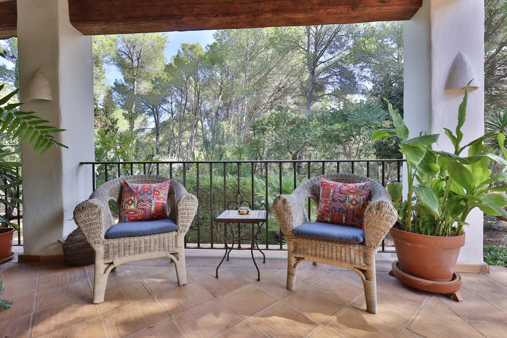 Terraza cubierta con dos sillones de mimbre y cojines azules, mesa auxiliar de forja, suelo de terracota y vistas a la exuberante vegetación natural. Terraza cubierta con dos sillones de mimbre y cojines azules, mesa auxiliar de forja, suelo de terracota y vistas a la exuberante vegetación natural.